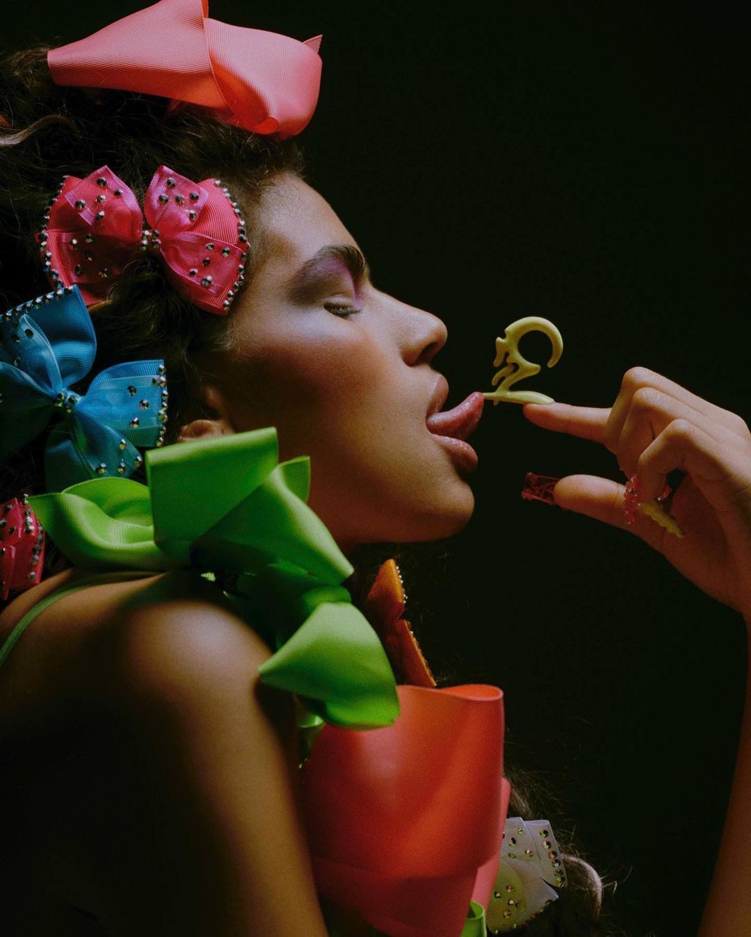 Woman with elaborate hair and makeup poses, tongue out, holding edible art. The image features bows, vibrant colors, and dark backdrop.