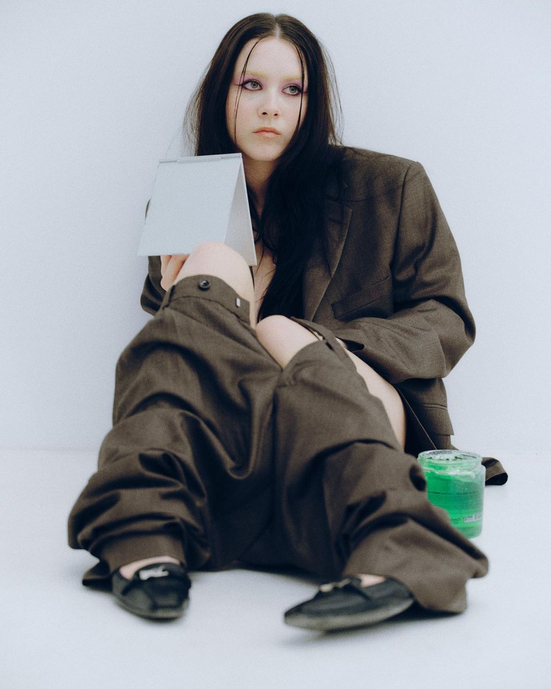 A young adult woman sits in a photo studio dressed in a brown suit with a mirror in her hand. Green liquid sits nearby.