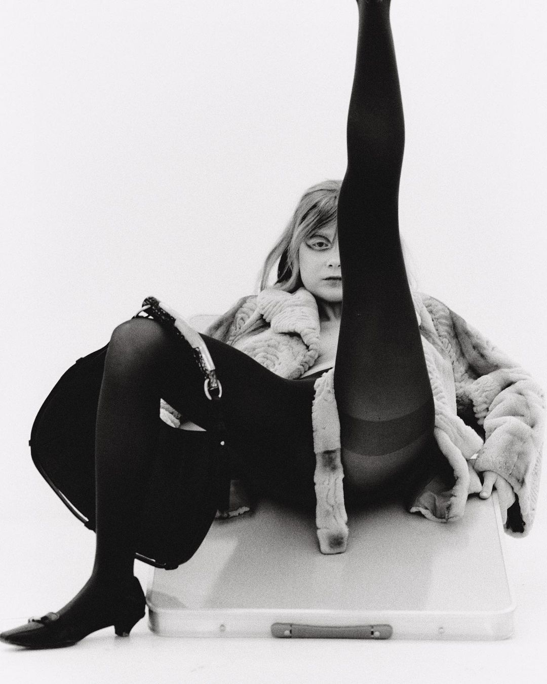 Black and white portrait of a woman in a fur coat, tights, and heels, with one leg raised vertically. She sits on a suitcase.