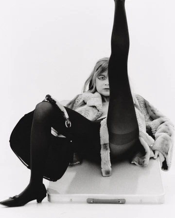 Black and white portrait of a woman in a fur coat, tights, and heels, with one leg raised vertically. She sits on a suitcase.
