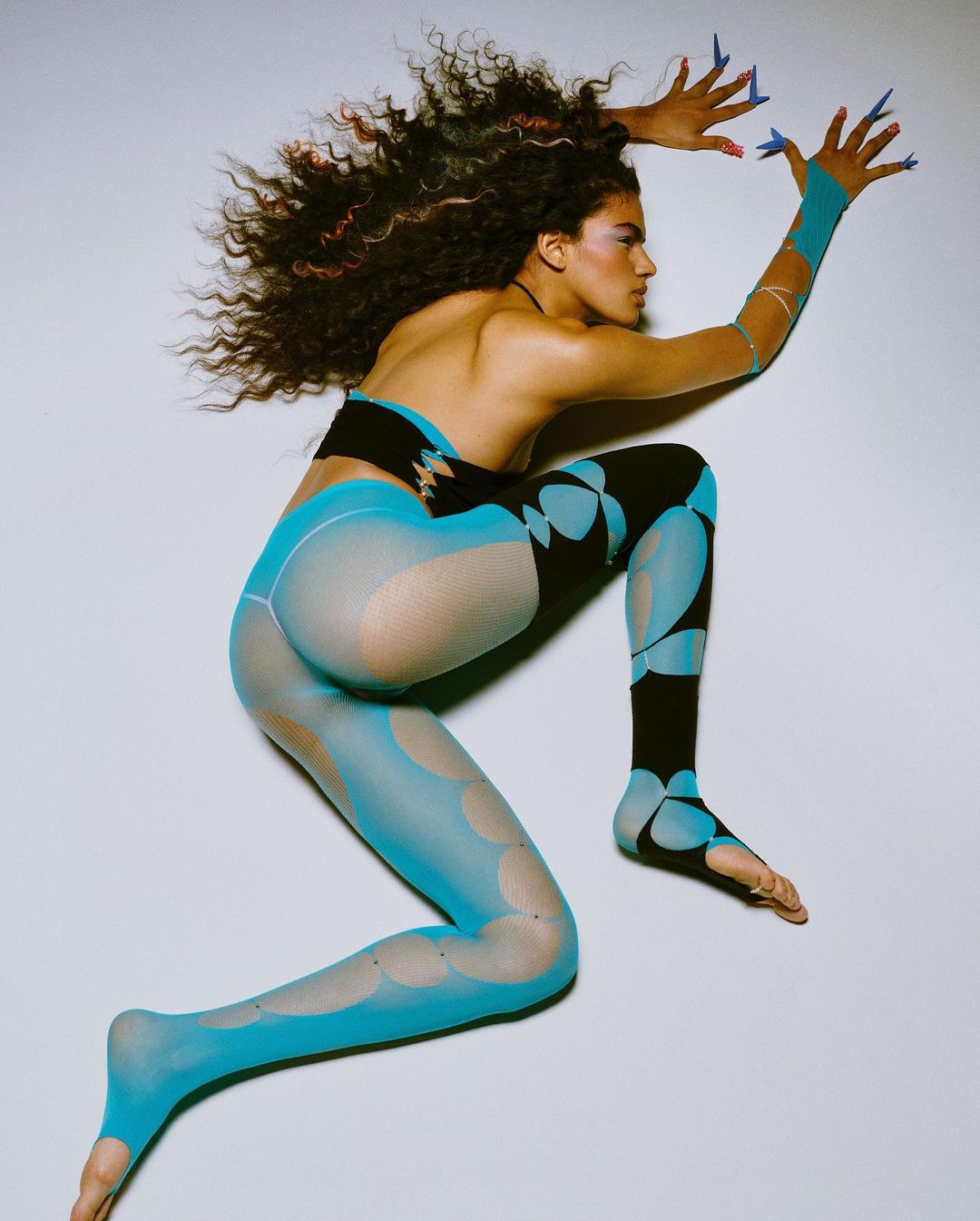 Woman in patterned tights and crop top poses on a white background, showcasing a fashion-forward and artistic studio portrait.