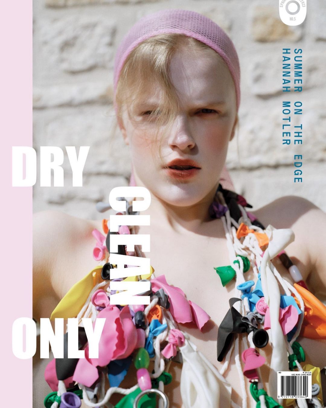 Magazine cover featuring a fair-skinned woman with a pink headscarf and a colorful garment of balloons against a neutral wall backdrop.