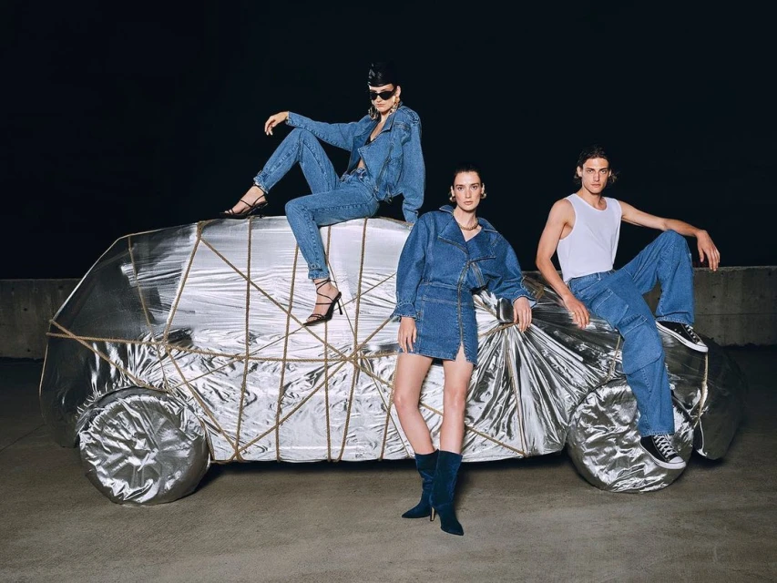 Three models in denim outfits pose on and around a silver foil-covered car, set against a dark background in a fashion photoshoot.