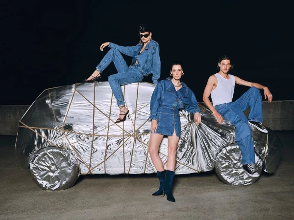 Three models in denim outfits pose on and around a silver foil-covered car, set against a dark background in a fashion photoshoot.