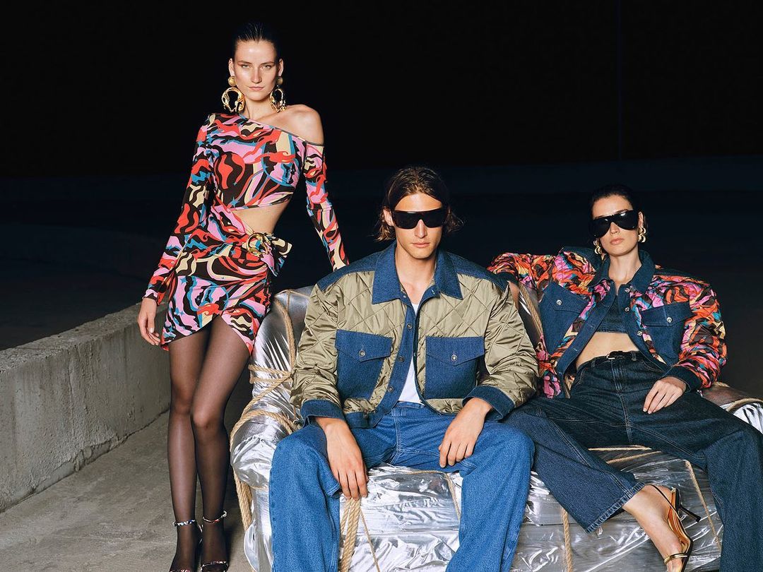 Three fashion models wearing colorful patterned clothing and sunglasses pose on a silver sofa in a dark outdoor setting.