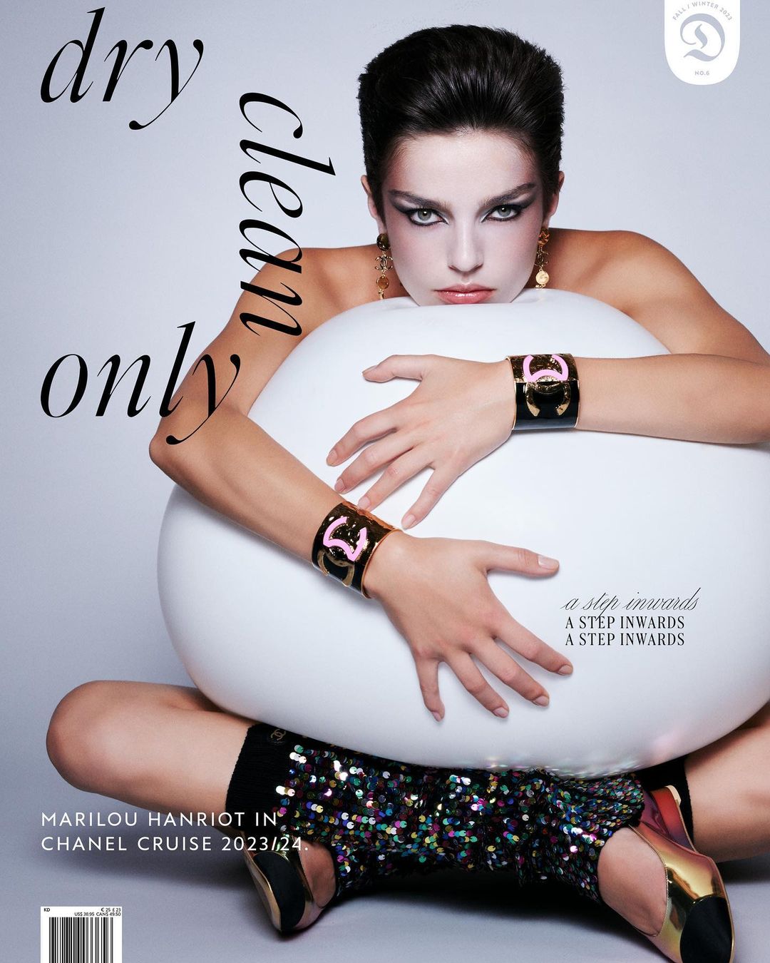 Fashion magazine cover featuring a model with dark hair, wearing jewelry and a sparkling dress, seated on a white round prop.