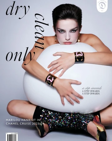 Model with dark hair and bold eye makeup hugging a large white sphere, wearing gold cuffs and sequin boots.