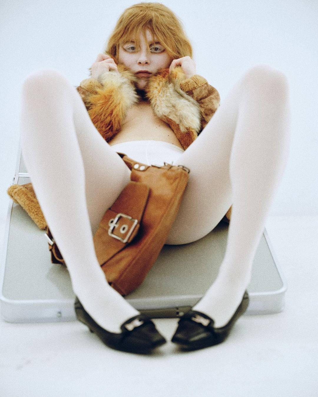 A young, light-skinned woman with ginger hair poses in a furry cropped jacket, white tights, and black flats, sitting in a white studio.