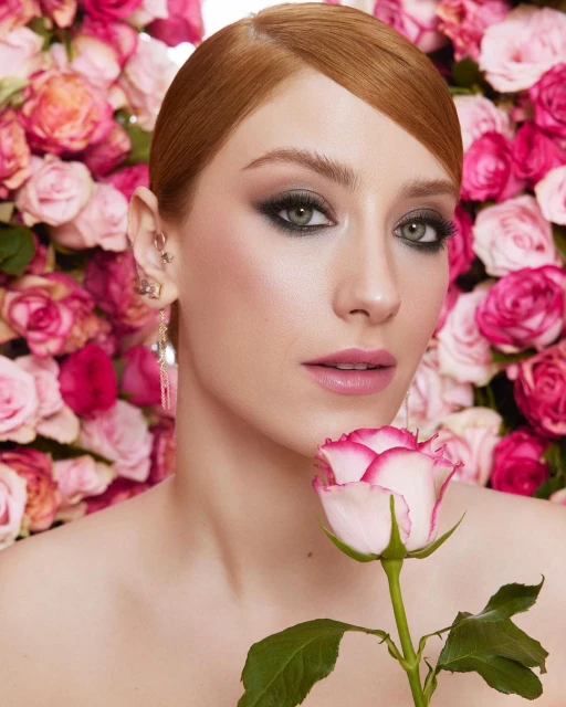 Close-up portrait of a woman with red hair, wearing makeup and jewelry, holding a rose, with a pink rose background.