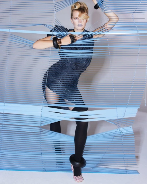 Fashionable woman poses in a short blue dress, black stockings, and pink shoes, framed by blue blinds, in a studio setting.
