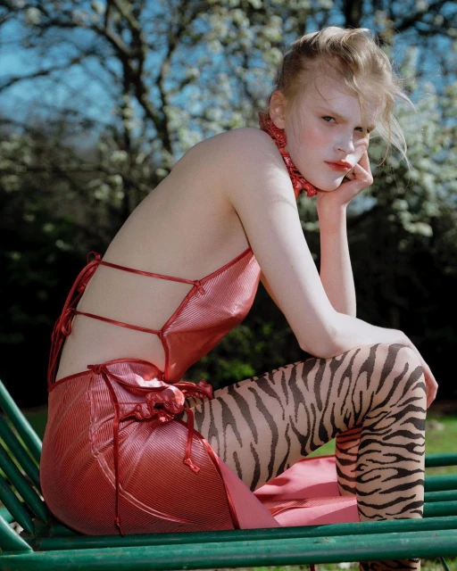 A young, fair-skinned woman in a red dress and tiger-print tights sits on a green bench against a backdrop of blooming trees and a blue sky.