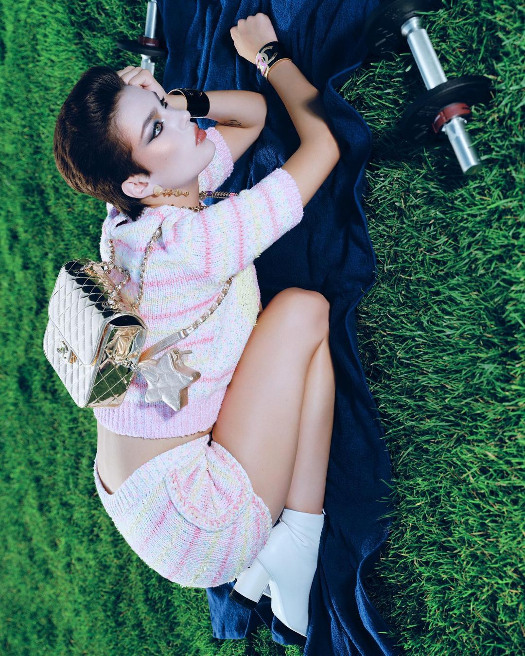 Woman in knitwear poses with dumbbells on grass, accessorized with Chanel bag and white boots, creating fitness-fashion blend.