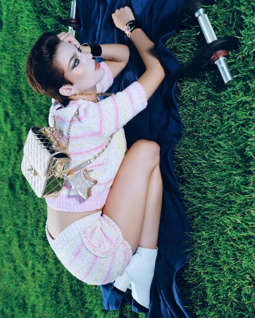 Woman in knitwear poses with dumbbells on grass, accessorized with Chanel bag and white boots, creating fitness-fashion blend.
