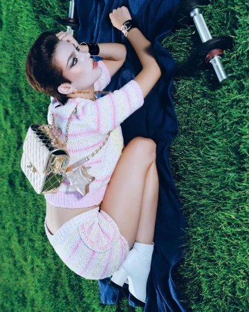 Woman in knitwear poses with dumbbells on grass, accessorized with Chanel bag and white boots, creating fitness-fashion blend.