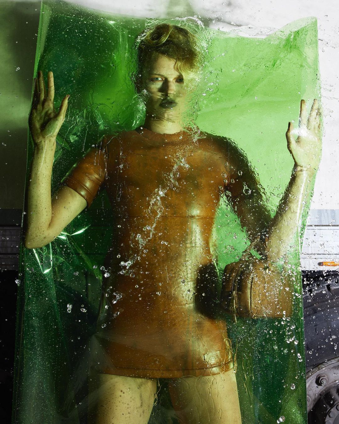 A person trapped in a sheet of transparent green plastic with water droplets, wearing a short, bronze-colored latex dress and wrist accessory.