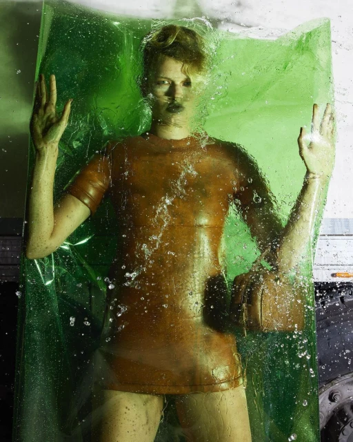 A person trapped in a sheet of transparent green plastic with water droplets, wearing a short, bronze-colored latex dress and wrist accessory.