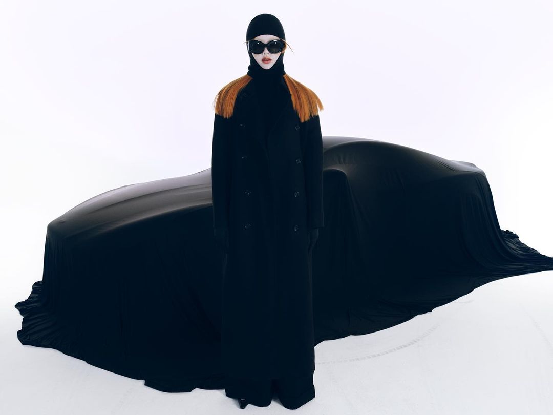 Fashion shot of a woman in a black coat, hat, and sunglasses standing in front of a car covered by a black tarp in a studio.
