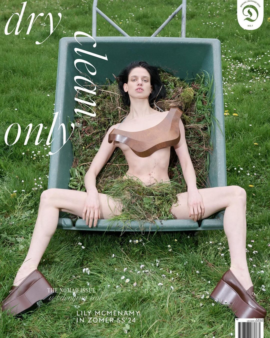 Magazine cover shot of a woman lying in a wheelbarrow filled with grass clippings wearing oversized shoes and a geometric wooden top in a lawn.