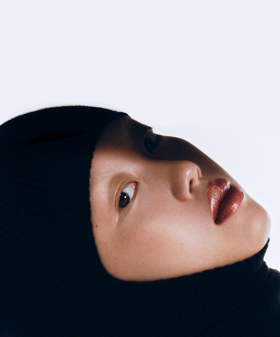 Close-up portrait of an adult woman with fair skin wearing a black hooded top, looking up toward the bright white background.