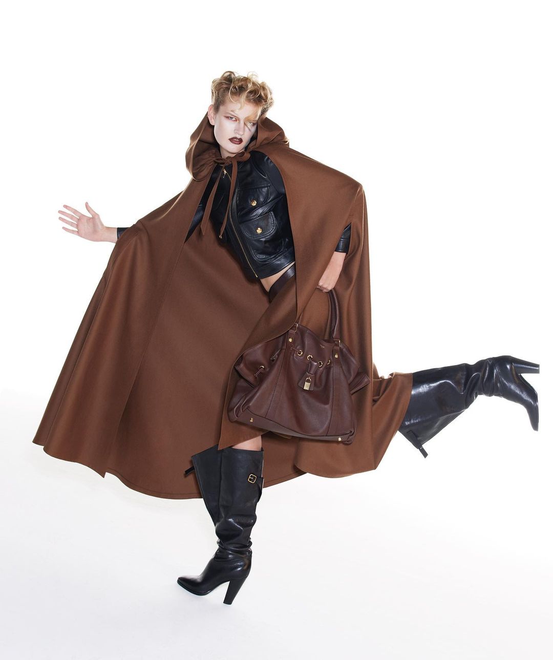 Fashion shot of a woman with blonde hair wearing a brown cape, black jacket, black boots, and holding a brown leather handbag against a white background.