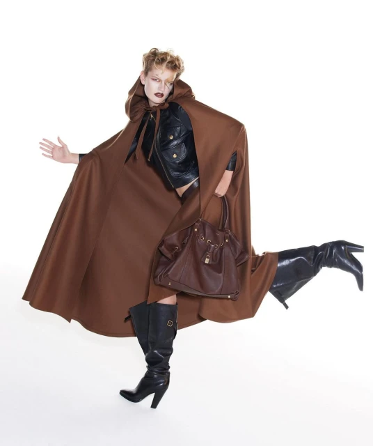 Fashion shot of a woman with blonde hair wearing a brown cape, black jacket, black boots, and holding a brown leather handbag against a white background.