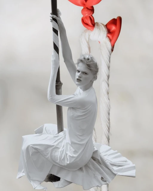 Monochrome fashion shot of an androgynous person in white dress, clinging to a striped pole under a large red-and-white rope knot.