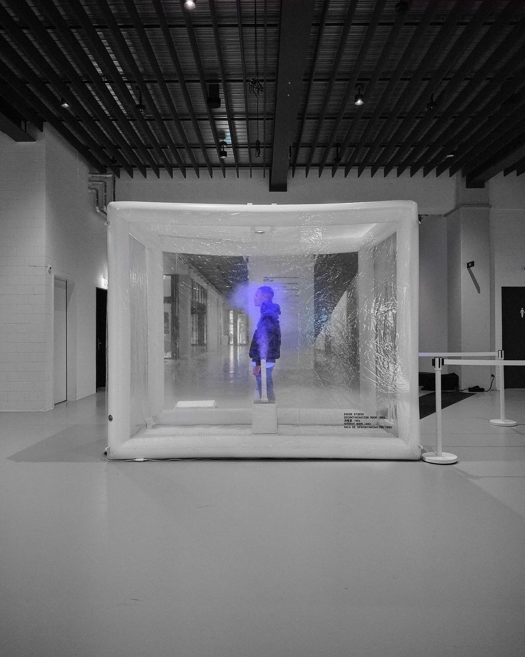 Abstract black and white shot featuring a person inside an illuminated, translucent cube in a large indoor space, creating a surreal visual.