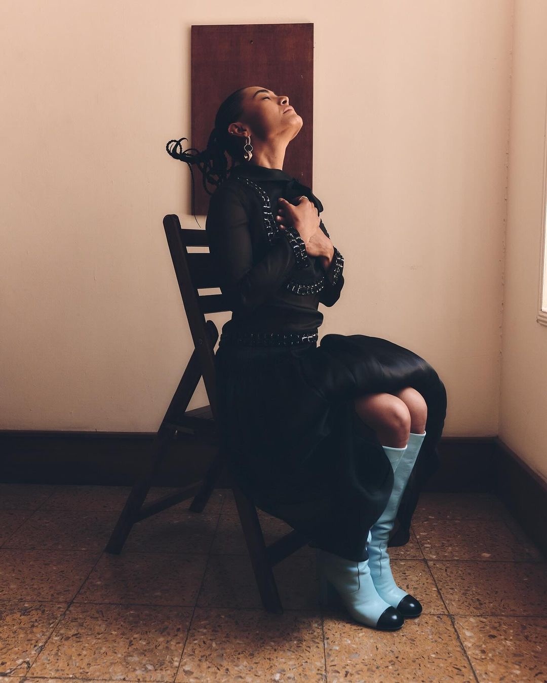 Adult female in stylish blue boots seated in wooden chair against a neutral background, posing with hands near her chest and eyes closed.