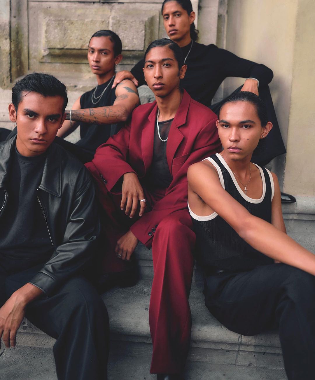 Five young adults are sitting on steps together, wearing fashionable clothing in a posed photograph, outside with natural light.
