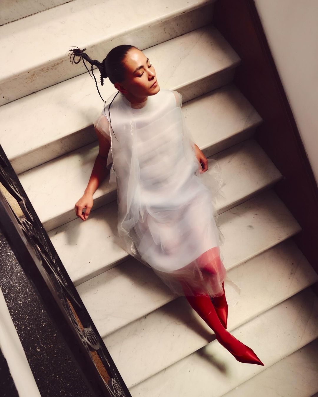 Woman in sheer white dress poses on marble stairs with red boots. Low angle view highlighting fashion and architectural details.