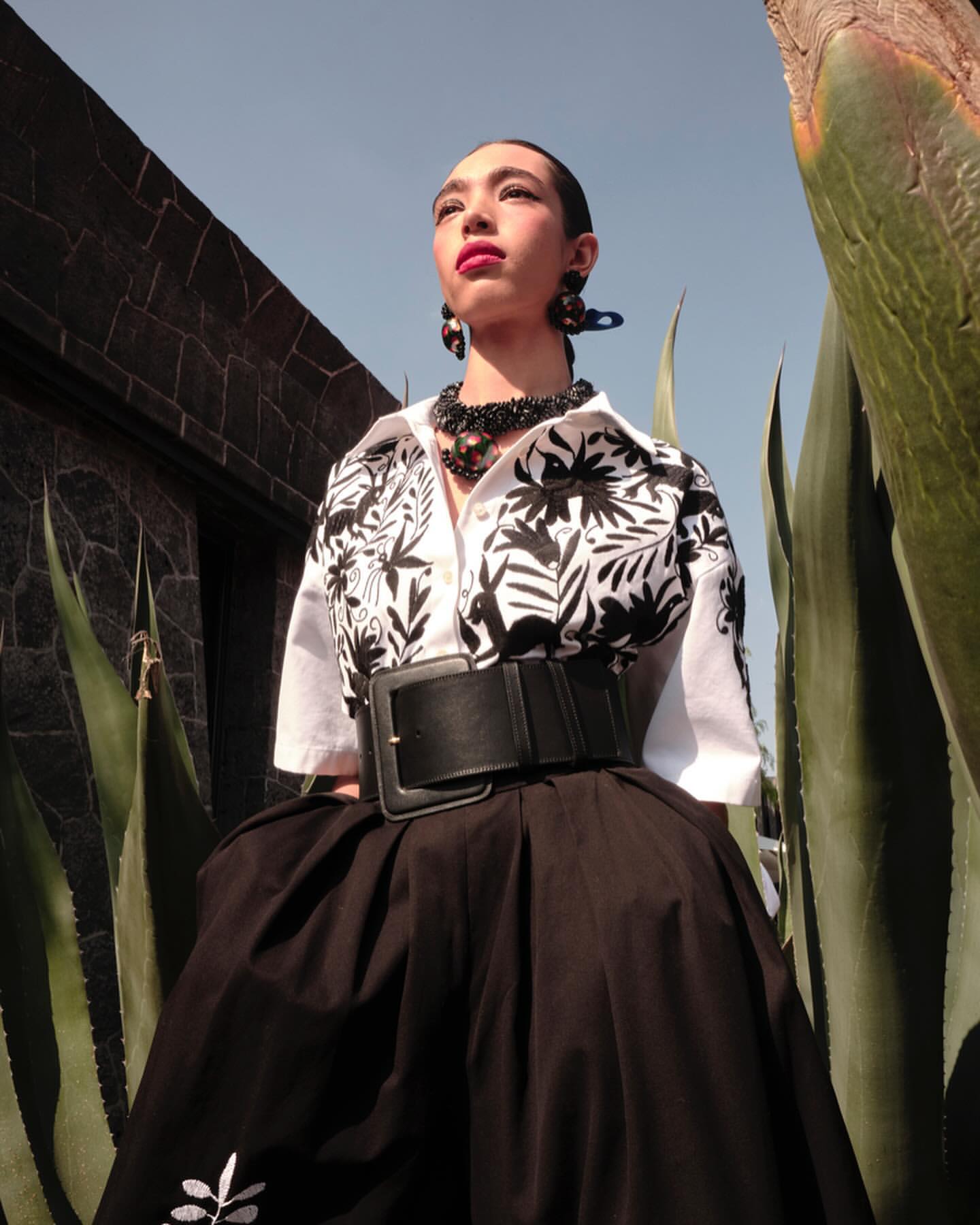 Model wearing white patterned shirt and black skirt with wide belt, framed between agave plants in New York.