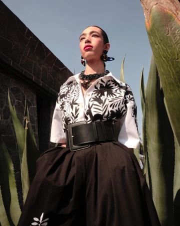 Model wearing white patterned shirt and black skirt with wide belt, framed between agave plants in New York.