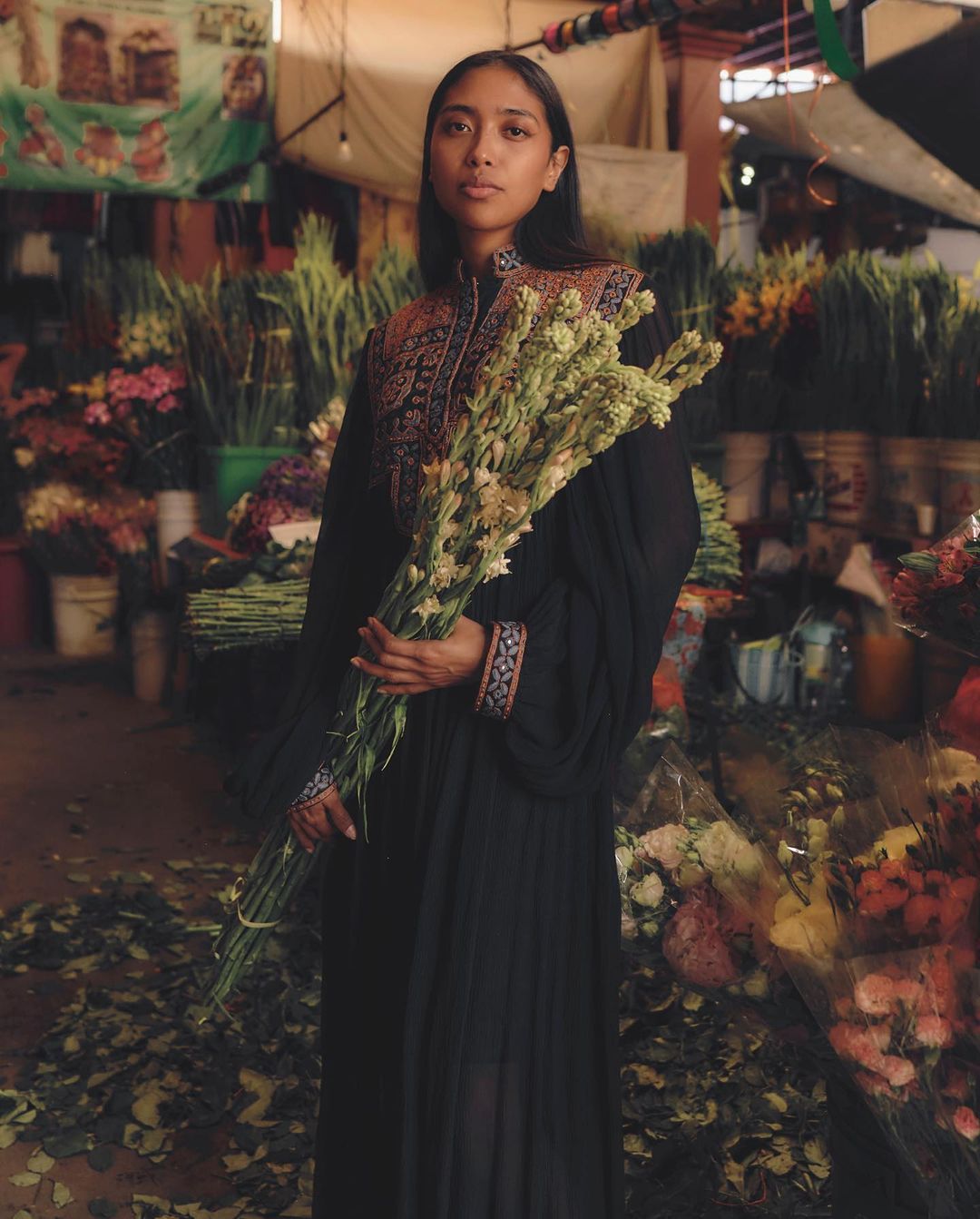 Woman in black dress holding white flowers in a flower market, with other flowers and market stalls in the background.