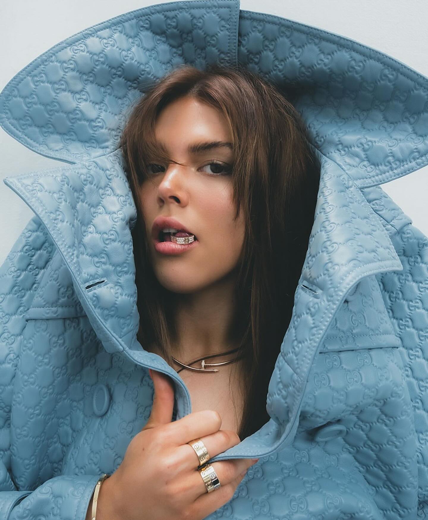 Portrait of a brunette adult female model wearing a blue textured jacket, gold rings, and a gold necklace indoors against a white background.