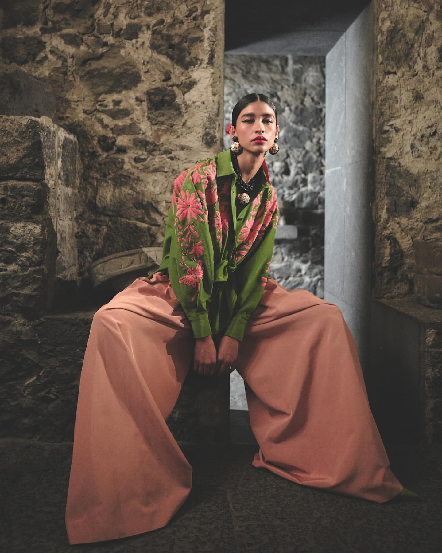 Fashion portrait of a woman in a green floral shirt and wide leg peach pants sitting against a stone wall in an indoor setting.