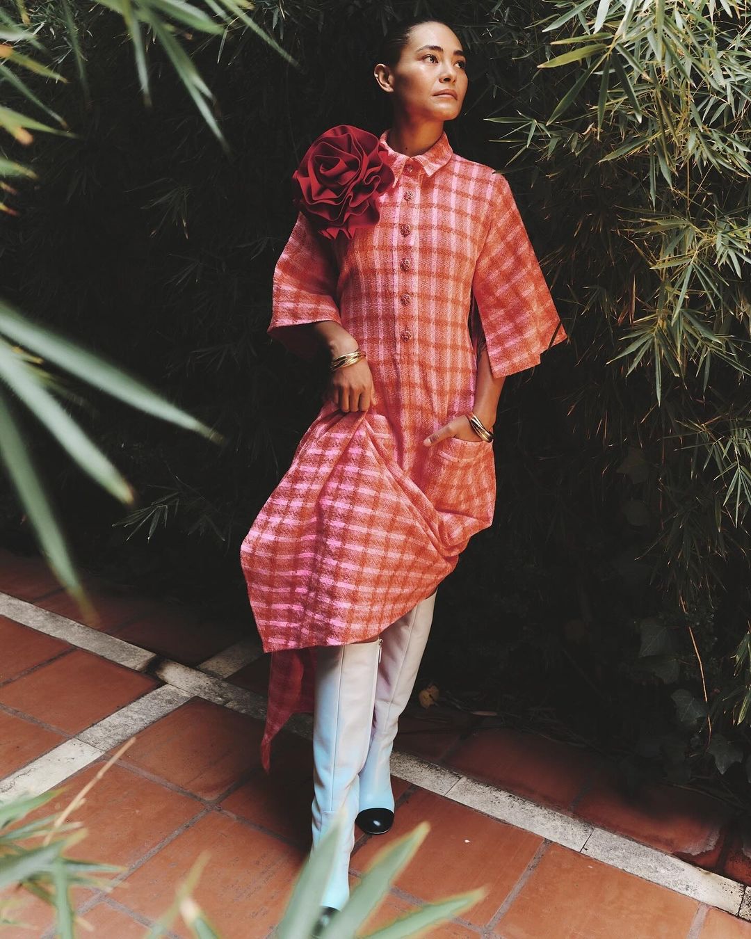 Woman poses outdoors in a pink plaid dress with a large flower detail, paired with blue boots on a terracotta tile patio.