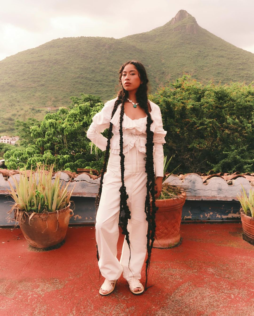 A woman in a white outfit poses outdoors on a rooftop with braided pigtails that reach her knees, with a mountain backdrop.