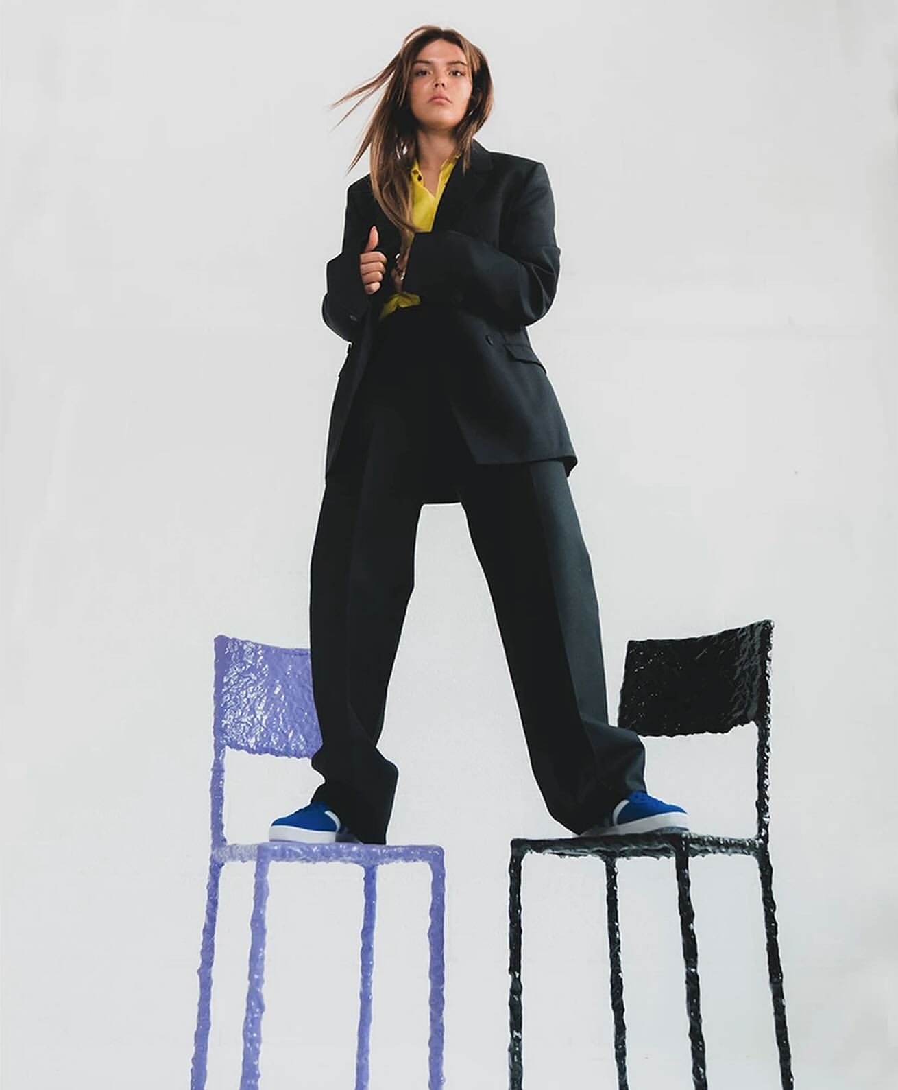 Stylish woman in oversized black suit and blue sneakers stands on painted chairs in a bright studio for fashion editorial photography.