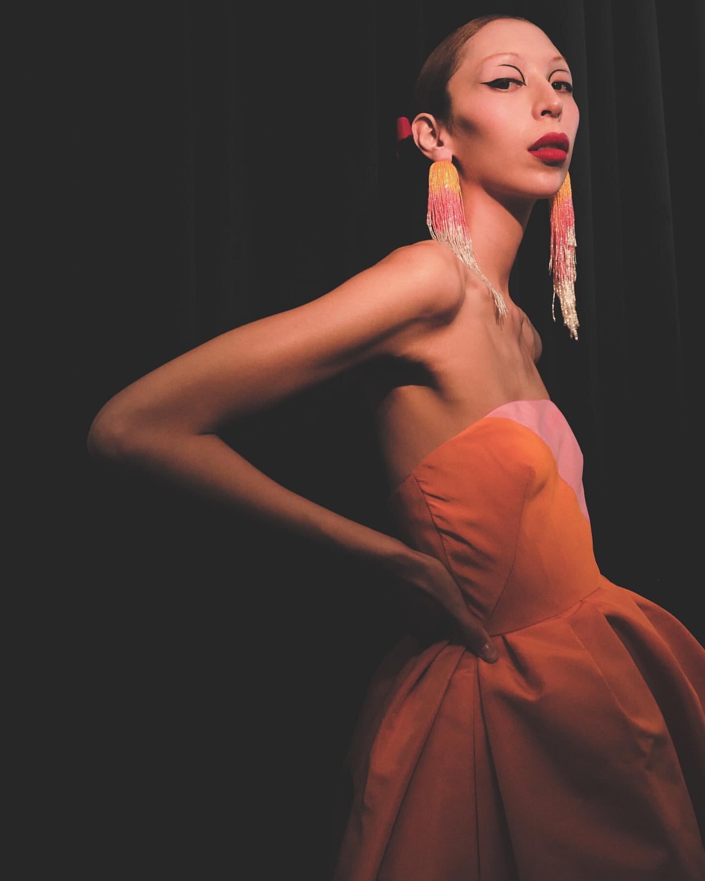Elegant adult female in a strapless peach and pink dress poses against a dark background with long beaded earrings and red lipstick.