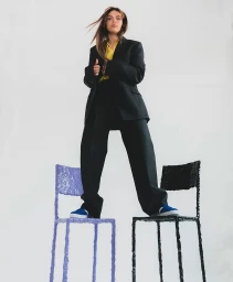 Model wearing a black suit jacket and trousers balanced with feet planted on two separate chairs against a plain white background.