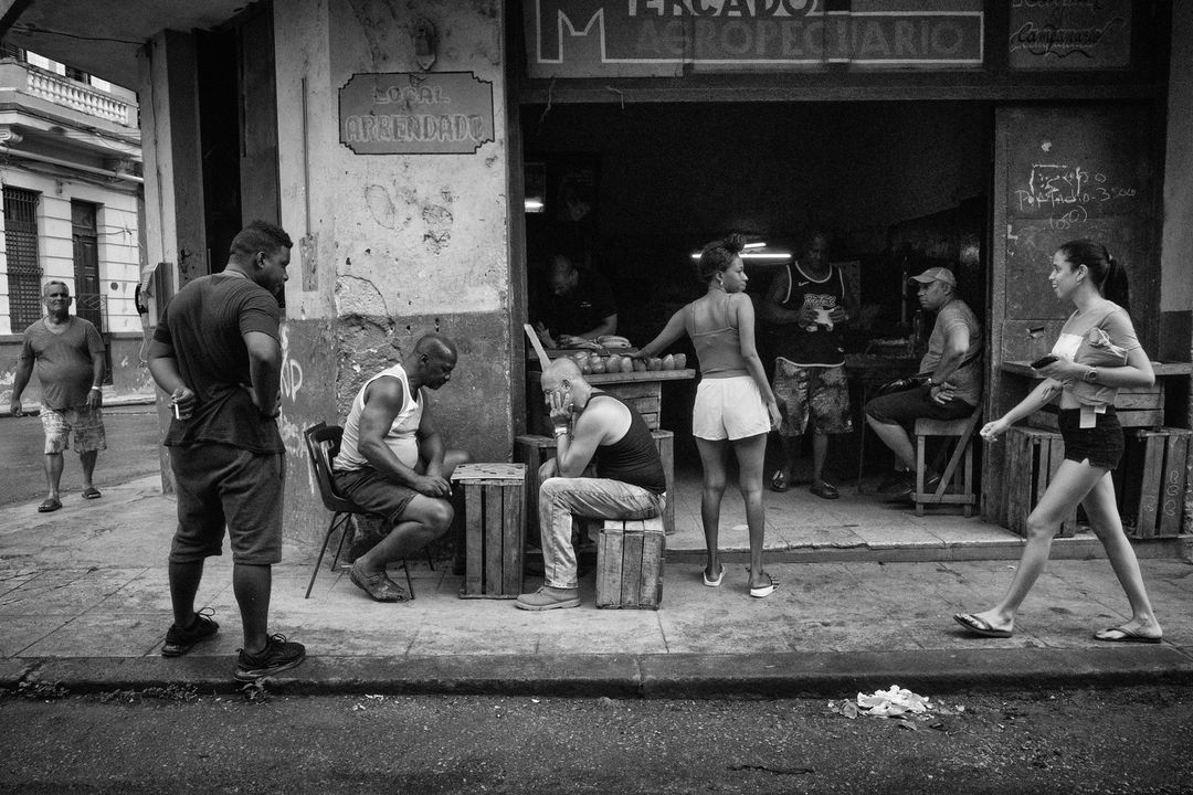 Monochrome street scene with locals and a woman walking by an open market, capturing candid daily life in an urban setting.