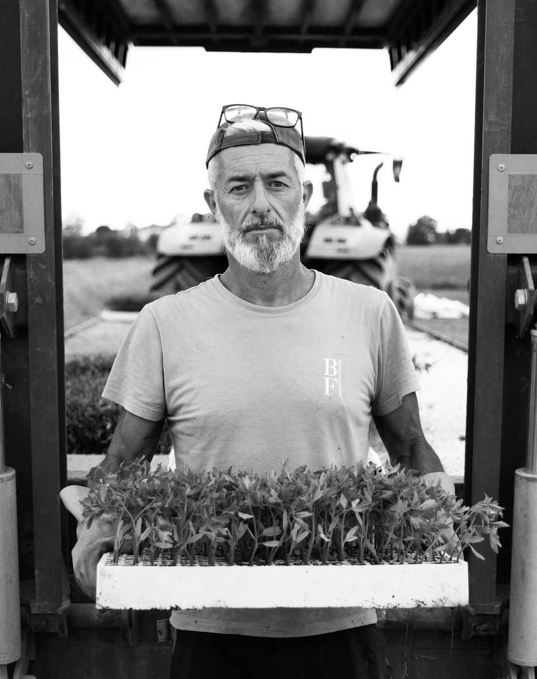 Monochrome portrait of a mature male farmer holding a tray of seedlings, standing outdoors in a field with a tractor visible in the background.