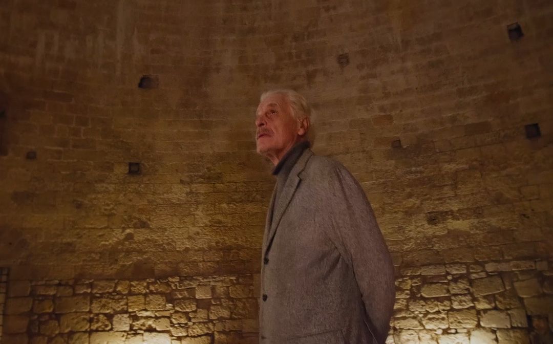 Elderly man in a grey coat stands inside a stone chamber with rounded walls. Lighting illuminates the textured surface.