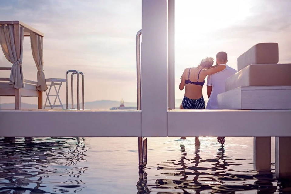 Couple enjoying sunset view from poolside lounge with turquoise water reflections and tranquil horizon. Summer vacation relaxation.