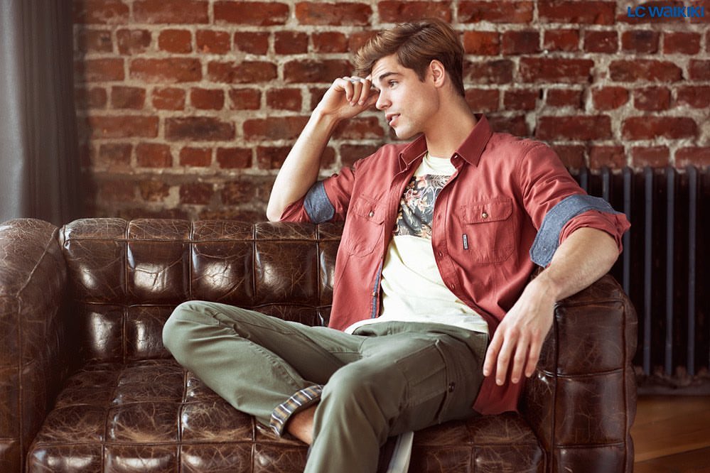 A young adult male sits relaxed on a distressed leather couch in a studio setting, wearing casual fashion with a brick wall backdrop.