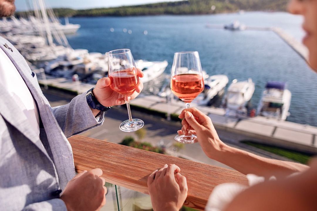 Elegant couple toasting with rosé wine glasses on a balcony overlooking a marina with boats, creating a romantic and celebratory atmosphere.