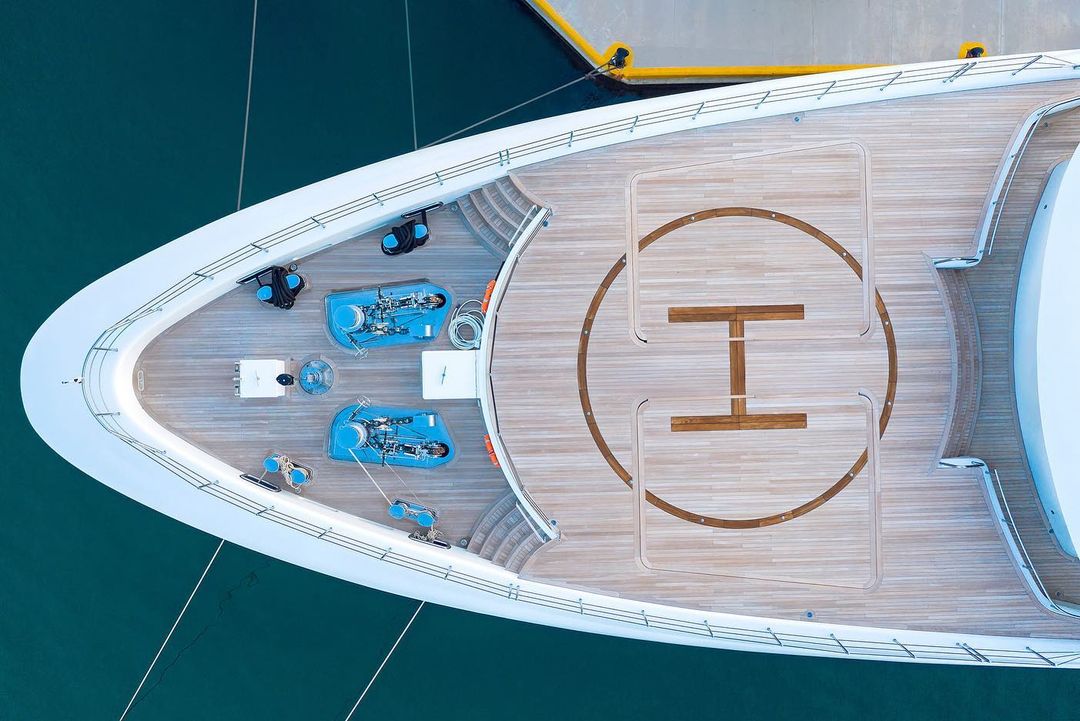 Aerial view of a luxury yacht deck featuring a helipad, seating areas, and equipment, docked in turquoise water on a sunny day.