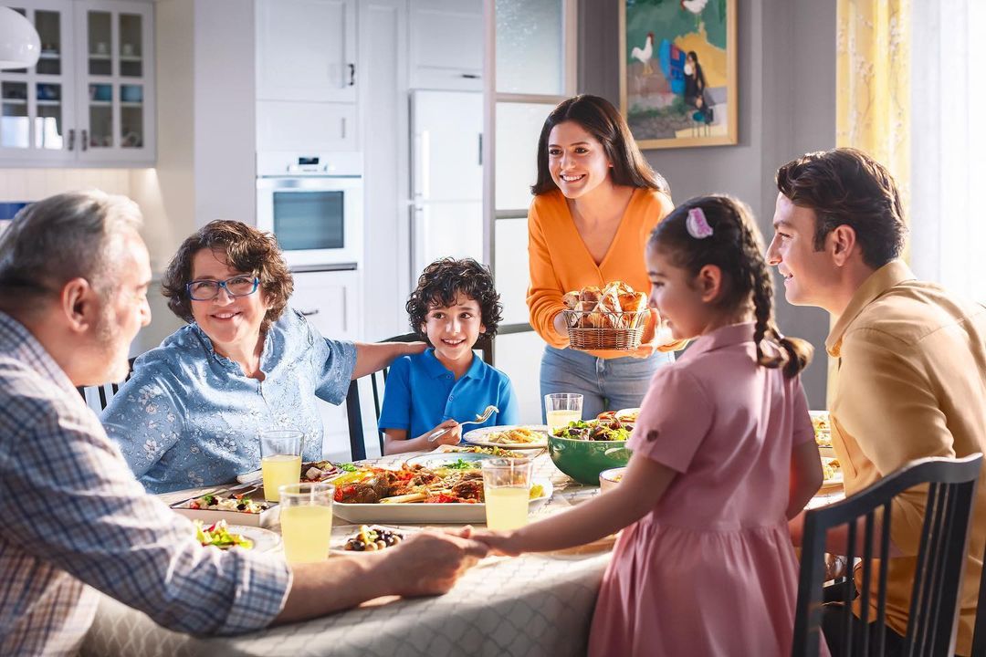 Multi-generational family enjoying meal at home dining table, including adults, children, serving dishes and bright modern interior.