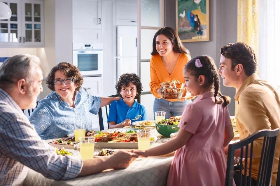 Multi-generational family enjoying meal at home dining table, including adults, children, serving dishes and bright modern interior.
