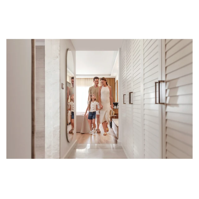 Family of three walks down a modern hallway with white walls and an oval mirror, showcasing a bright and clean hotel or home interior.